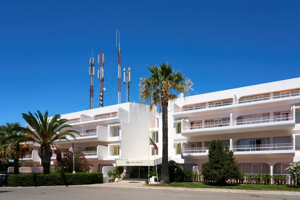 White hotel building with palm trees and communication towers against a clear blue sky.