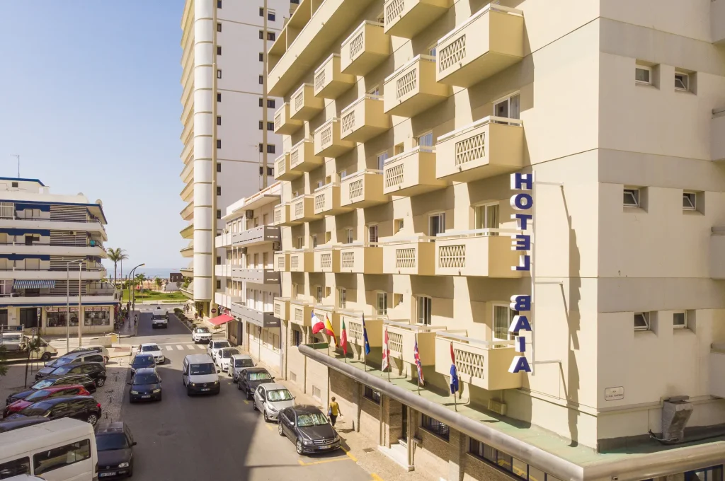 Street view of Hotel Baía with balconies, flags, parked cars, and nearby buildings under a clear sky.
