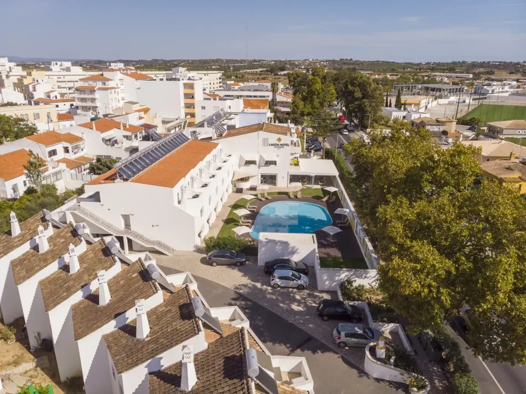 Aerial view of a hotel with a pool, surrounded by white buildings and trees in a residential area.
