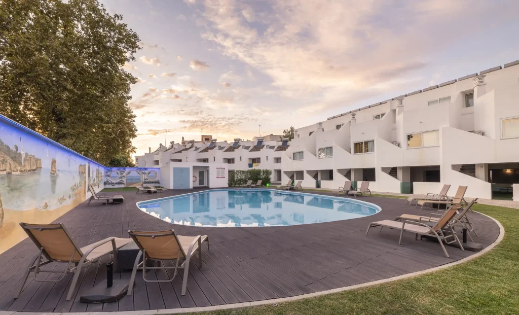 Outdoor pool with lounge chairs, mural, and white buildings at sunset.