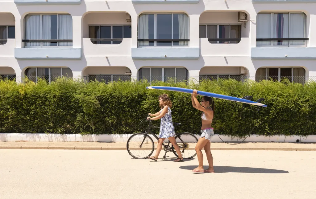 Woman walks with surfboard, another rides a bike. Background of green shrubs and white building. Sunny day.
