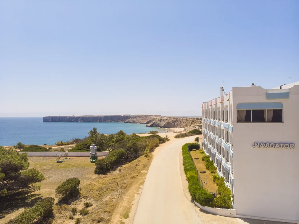 Coastal road by a white building labeled "Navigator," with a view of the ocean and distant cliffs under a clear sky.