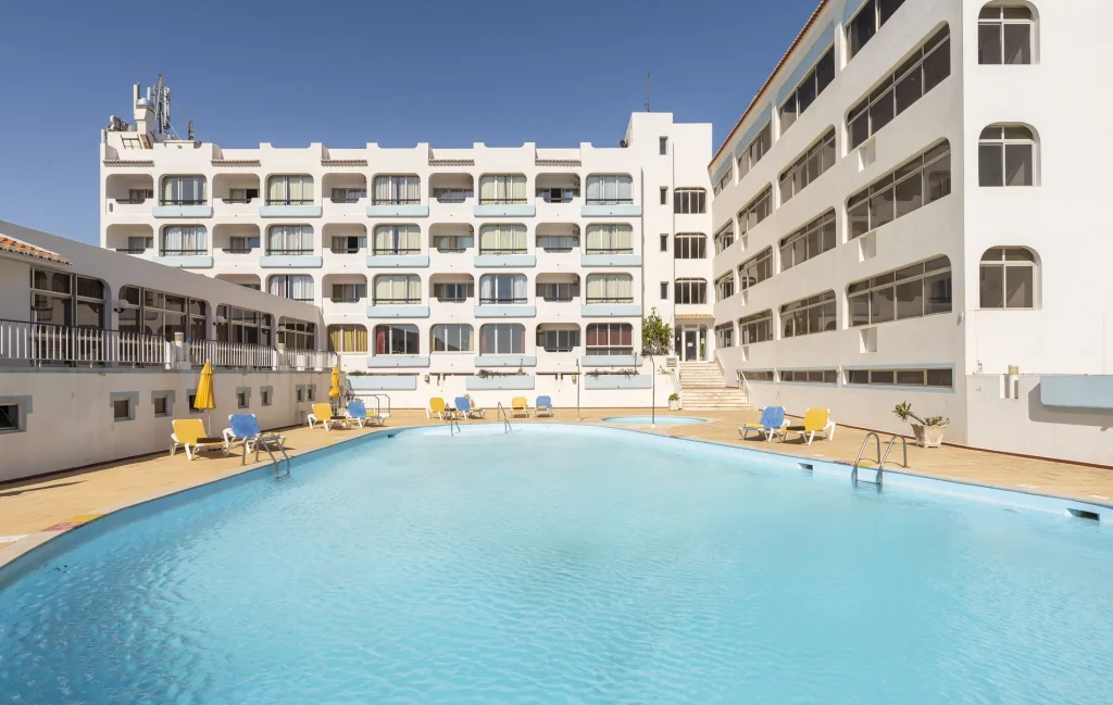 Outdoor pool with loungers, framed by a white hotel under a clear blue sky.