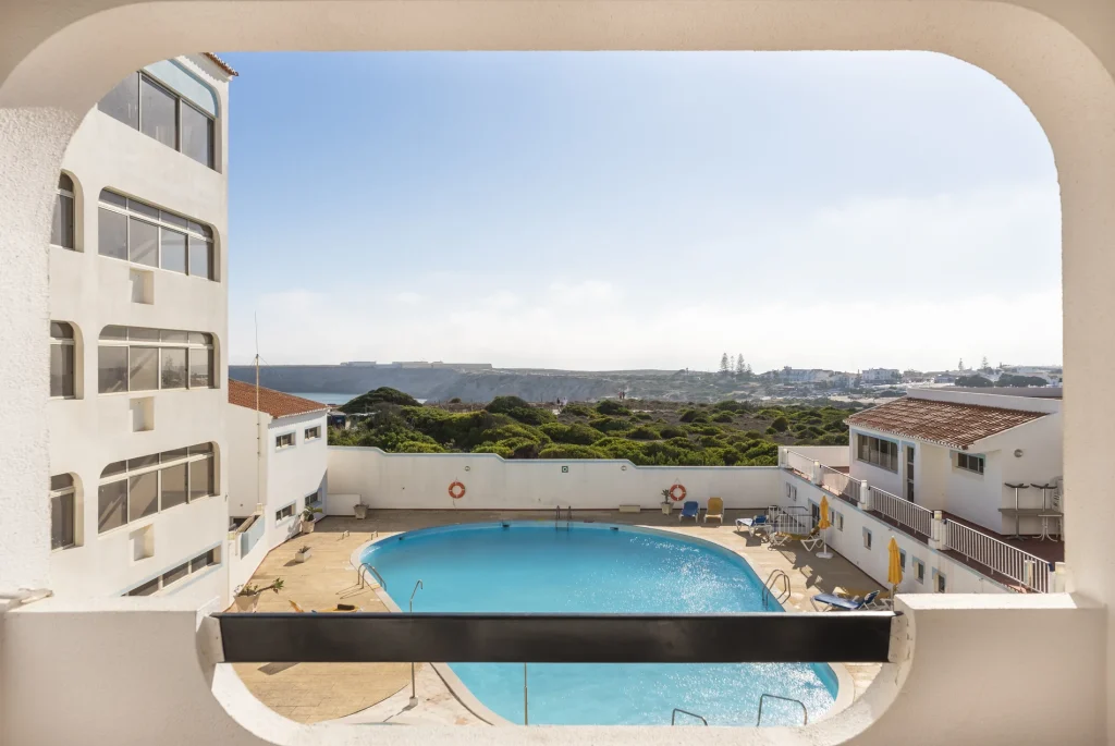 View of a swimming pool surrounded by white buildings, with greenery and distant hills under a clear blue sky.