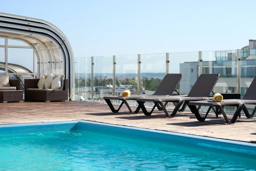 Rooftop pool with loungers, rolled towels, and cushioned seating under an open sky, surrounded by a glass barrier.