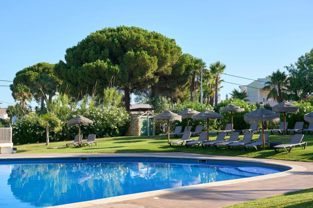 Poolside with loungers and parasols, surrounded by green trees and shrubs under a clear blue sky.