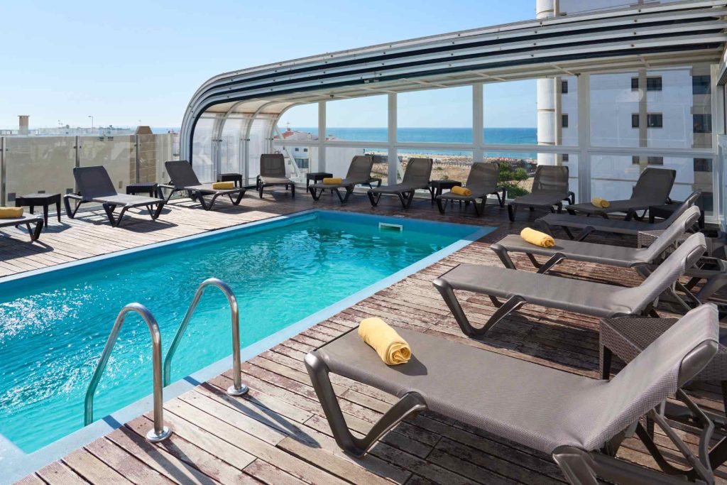 Rooftop pool with lounge chairs and sea view, surrounded by clear glass panels and sunlit sky.