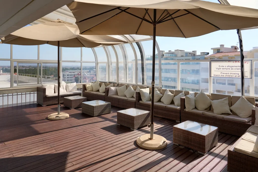 Rooftop terrace with wicker seating, cushions, glass tables, and large umbrellas, overlooking city buildings.