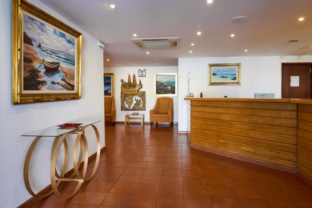 Hotel reception area with wood desk, paintings, and decorative chairs on tiled floor.