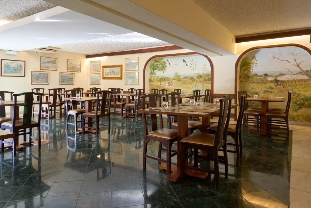 Spacious restaurant with wooden tables and chairs, wall art, and scenic murals, featuring glossy tiled flooring.