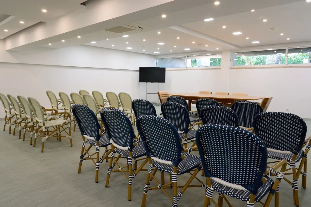 Empty conference room with rows of woven chairs facing a screen and table, under a ceiling with spotlights.