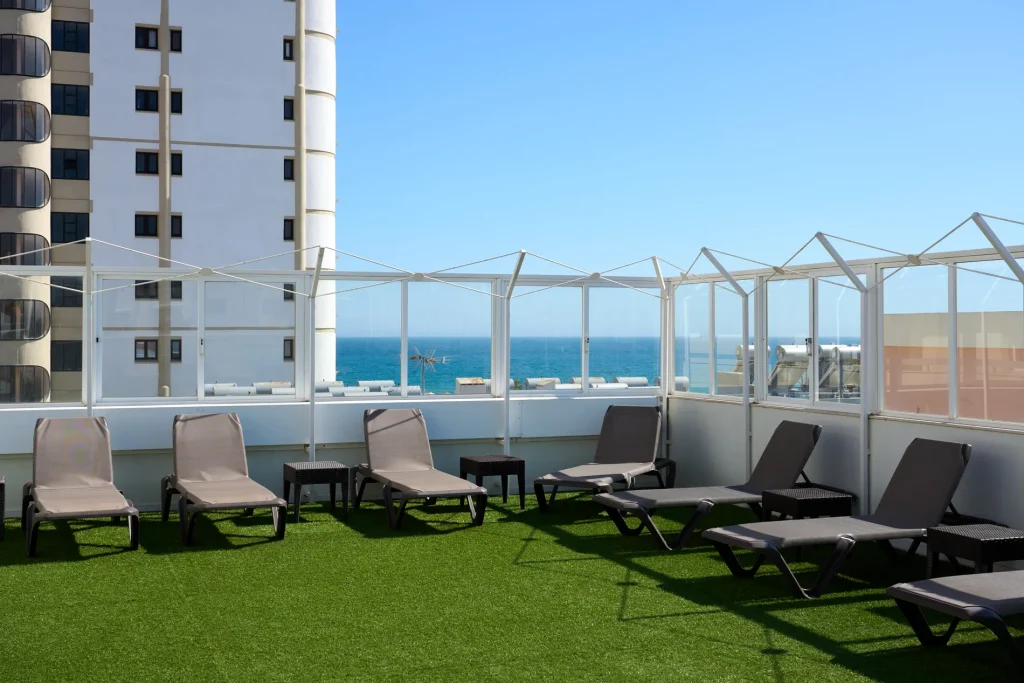Rooftop patio with lounge chairs, artificial grass, ocean view, clear blue sky, and neighboring buildings.