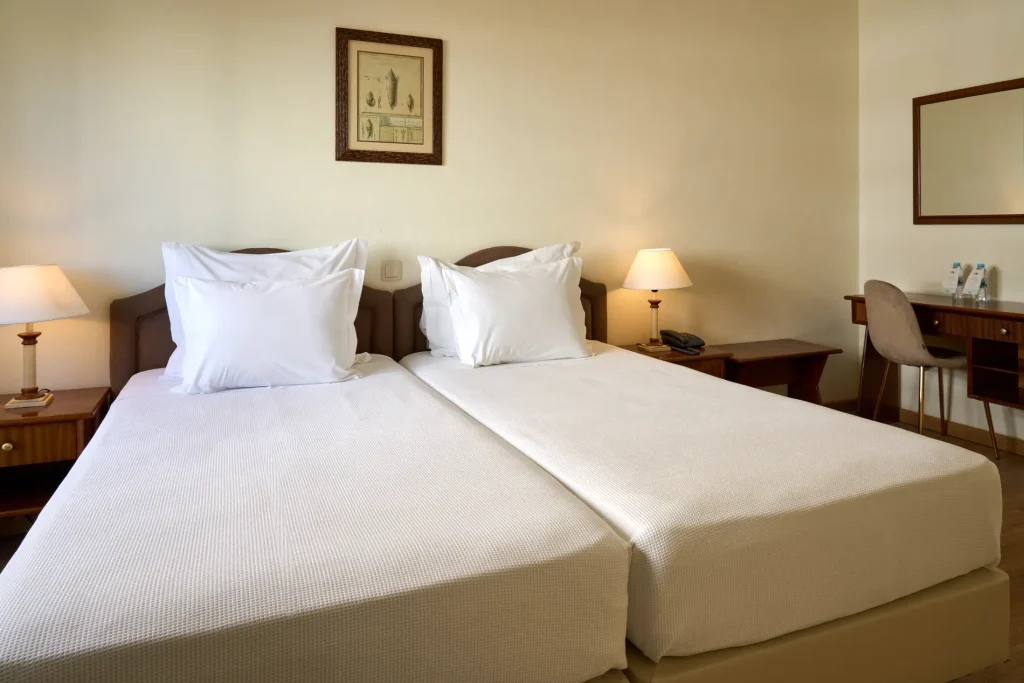 Hotel room with twin beds, white bedding, bedside tables with lamps, a desk with a chair, and a painting on the wall.