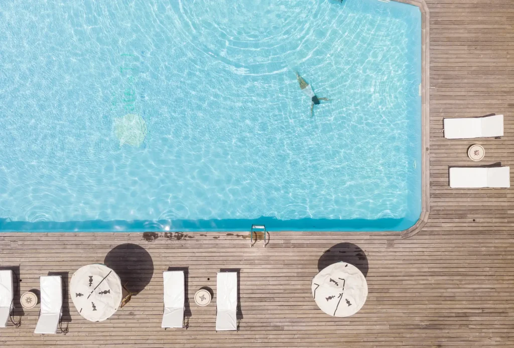 Aerial view of a swimming pool with a swimmer, surrounded by a wooden deck, loungers, and white umbrellas.