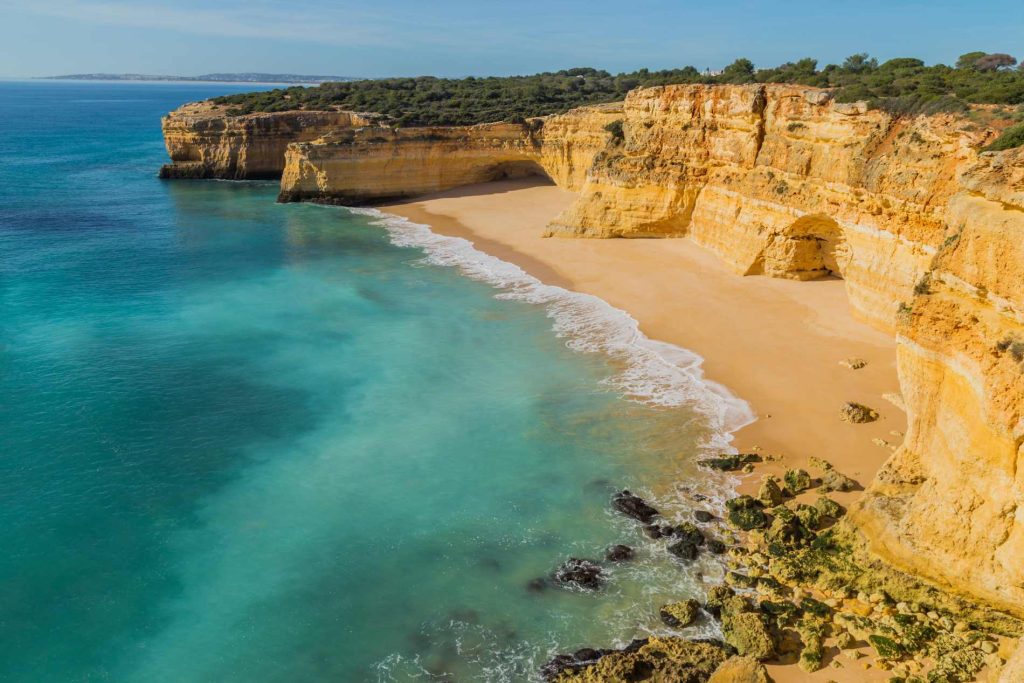 A secluded beach with turquoise water, surrounded by yellow cliffs under a clear blue sky.
