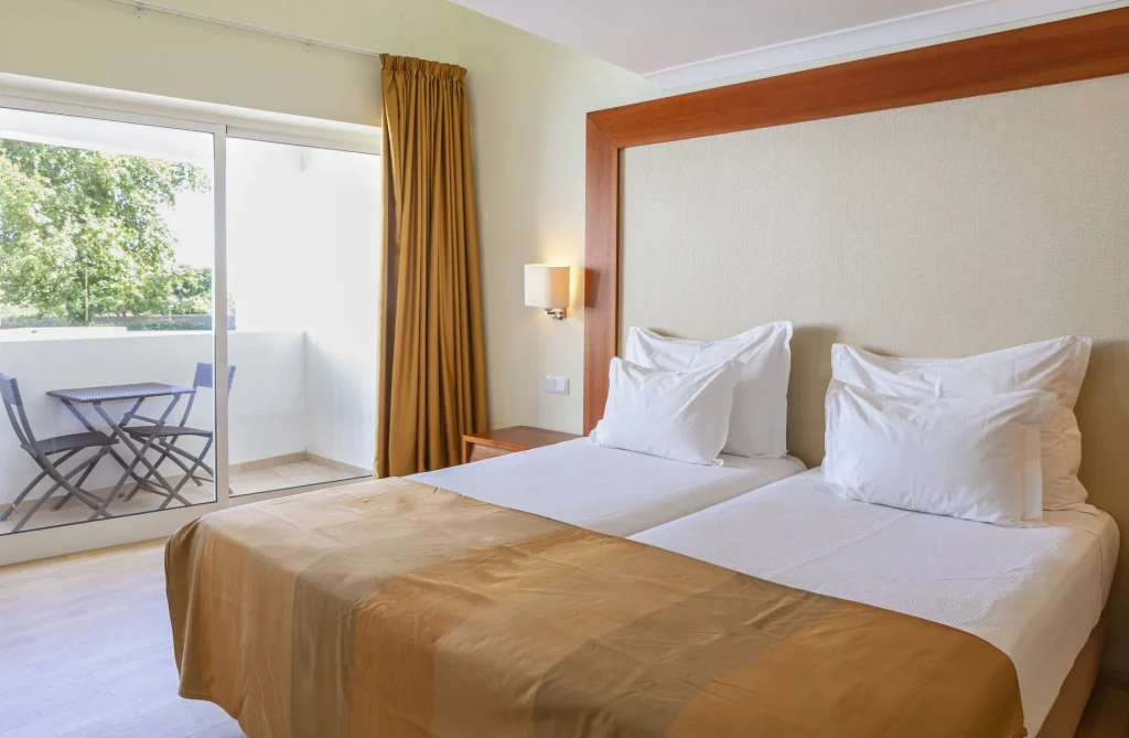 Hotel room with twin beds, beige headboard, sliding glass door to a balcony with table and chairs, and mustard curtains.