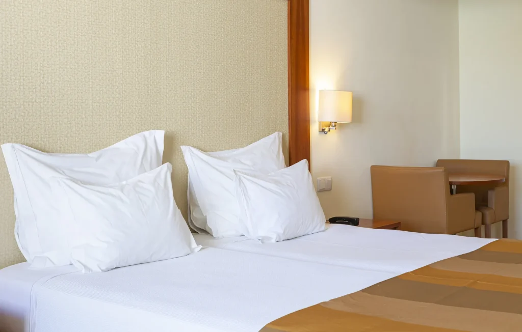Bed with white pillows and brown bedspread, wall lamp on, and a small table with two chairs in a cozy room.