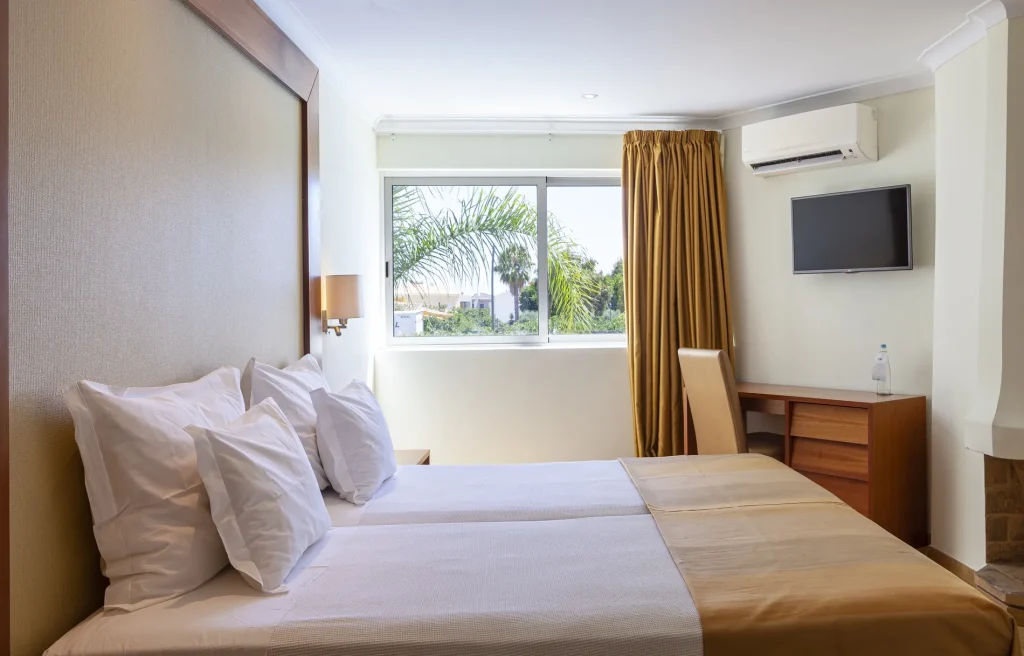 Hotel room with a double bed, pillows, a window with a palm view, a TV, desk, and air conditioning unit.