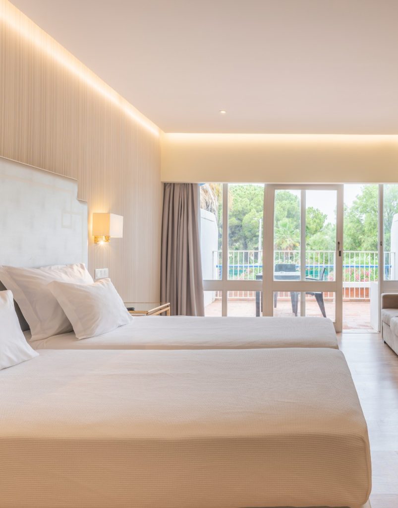 Hotel room with two beds, neutral decor, wall lamps, and a glass door opening to a balcony with a leafy view.