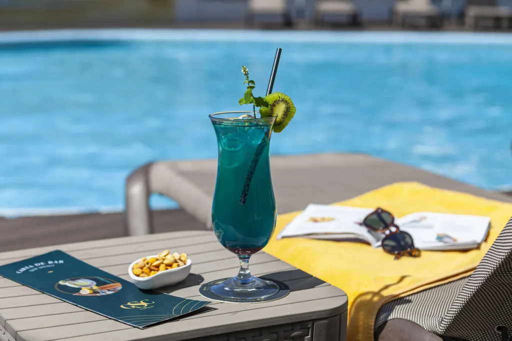 Blue cocktail by the pool with a kiwi garnish, nuts in a bowl, sunglasses, and a magazine on a yellow towel.