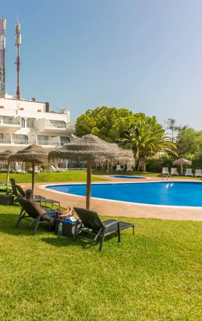 Man relaxing by a pool under a thatched umbrella, with a hotel and trees in the background.