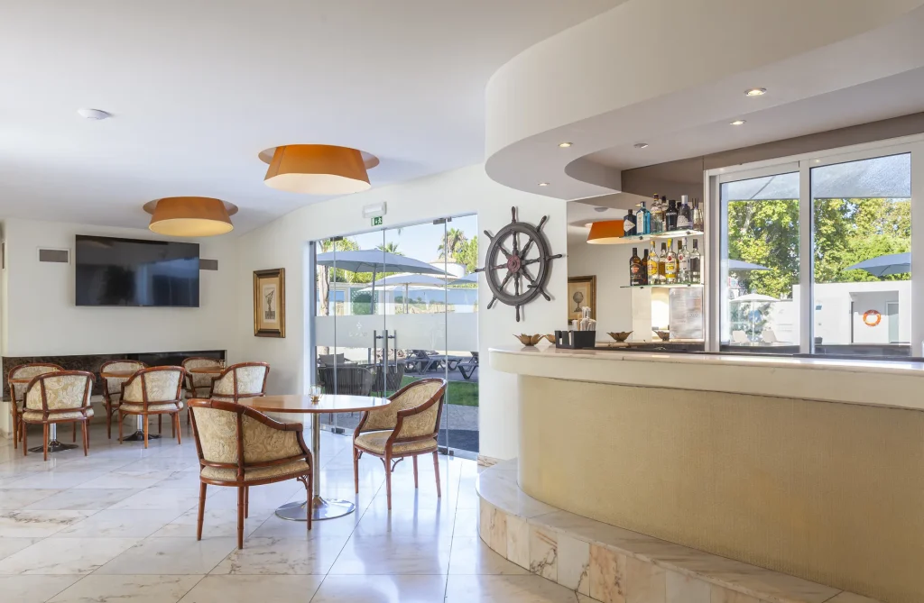 Modern hotel lounge with bar, nautical decor, overhead lights, glass doors, seating area, and garden view outside.