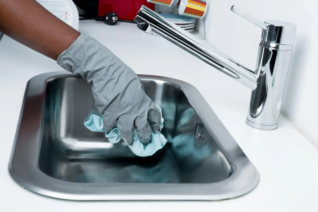 A gloved hand cleaning a stainless steel sink with a cloth next to chrome faucet in a kitchen.