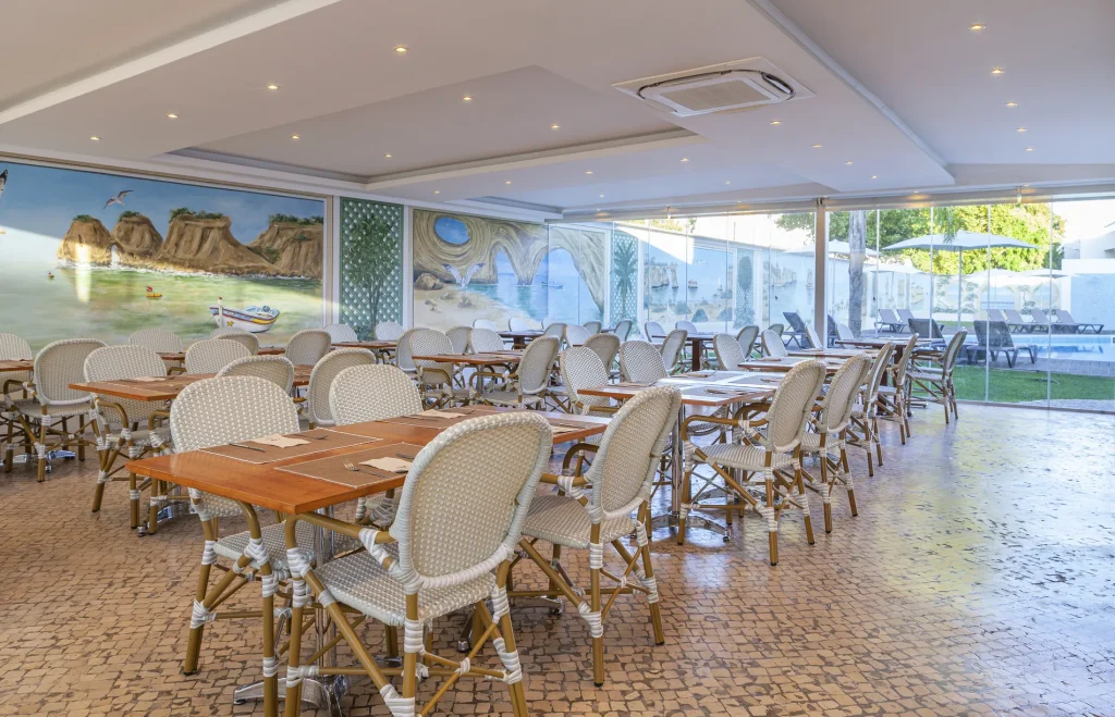 Bright restaurant interior with wicker chairs, wooden tables, and a mural of a coastal scene on the wall.