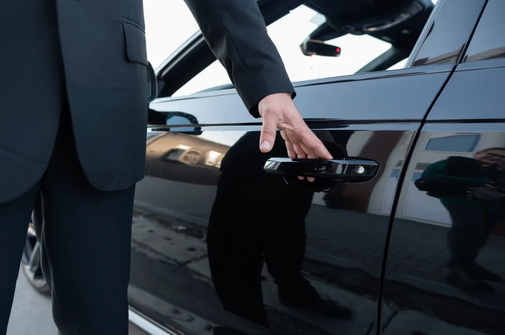 Person in a black suit opening a car door handle, with reflections visible on the car's surface.