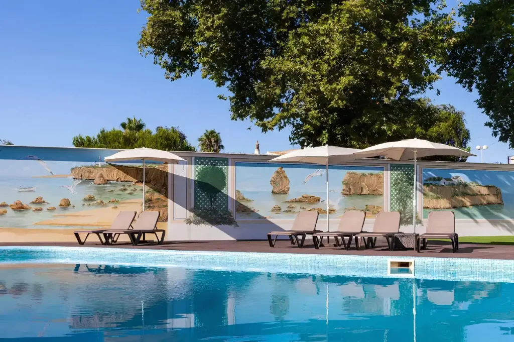 Poolside chairs under umbrellas, large beach mural backdrop, clear blue water, and sunny day with green trees.