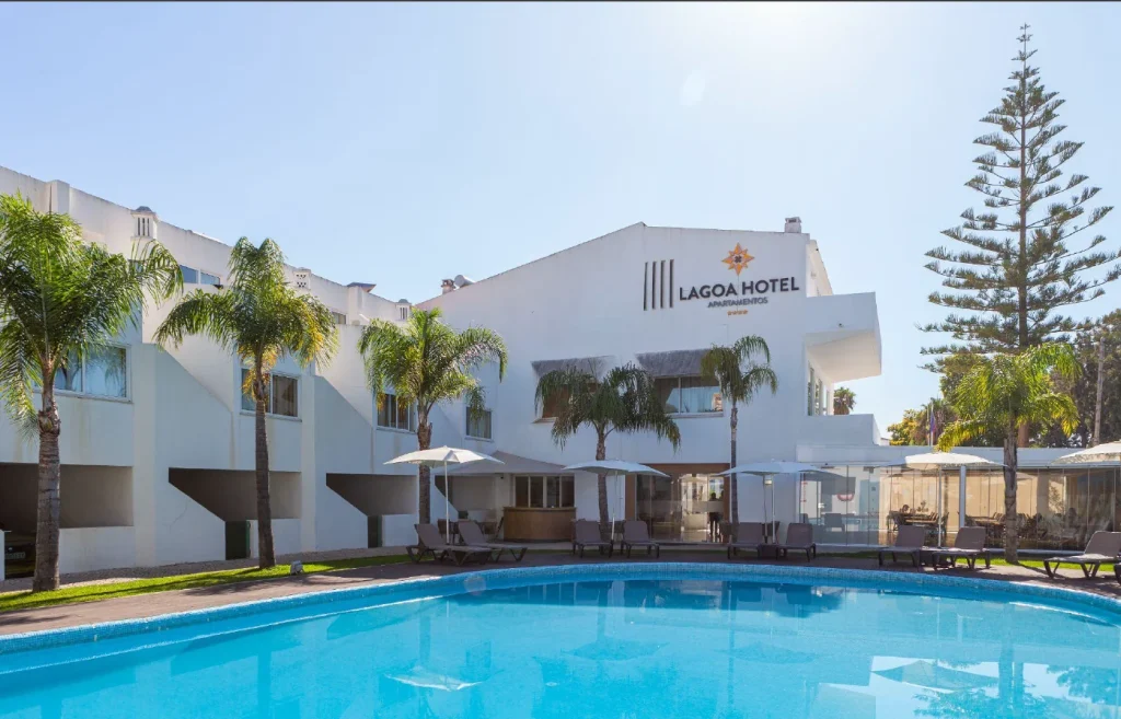 White hotel building with palm trees, sun loungers, and umbrellas around a blue pool under a clear sky.