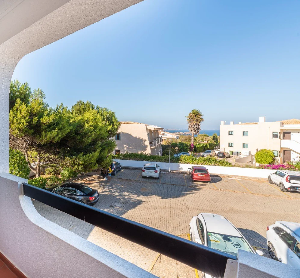View from a balcony overlooking a sunny residential area with parked cars, trees, and distant ocean.