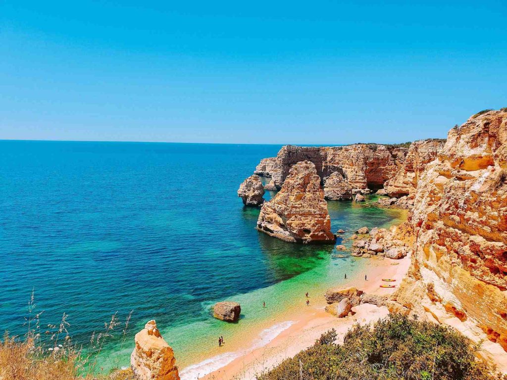 Rocky coastline with clear blue water and sandy beach, people swimming and relaxing on a sunny day.