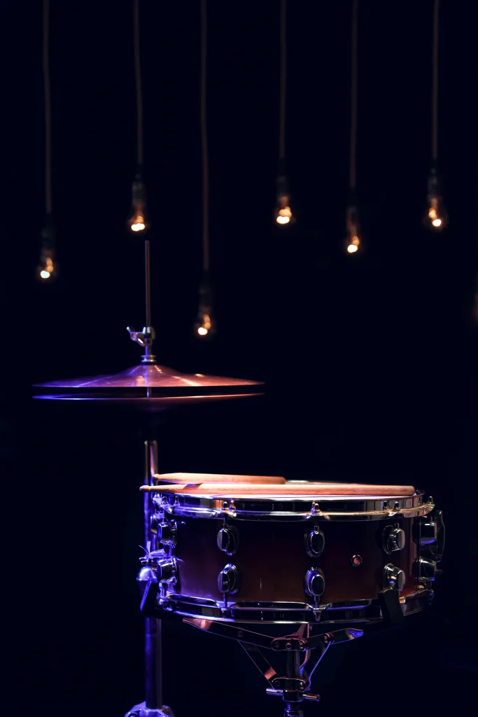 Snare drum with drumsticks and cymbal, under warm hanging lights on a dark stage.