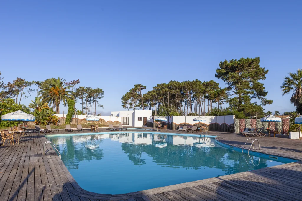 Outdoor pool with wooden deck, lounge chairs, and umbrellas surrounded by trees under a clear blue sky.