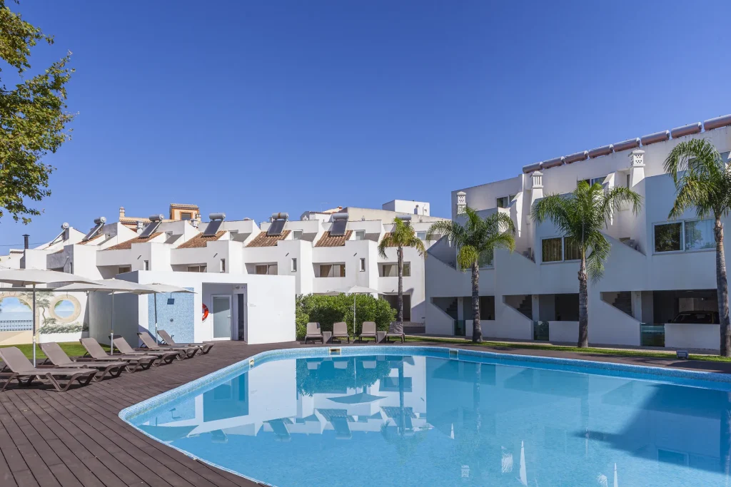 Modern white building with palm trees, a clear blue swimming pool, and lounge chairs under umbrellas on a sunny day.