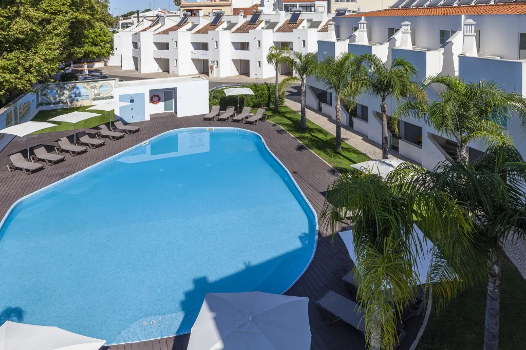Outdoor pool surrounded by lounge chairs, parasols, and palm trees, with white buildings in the background.
