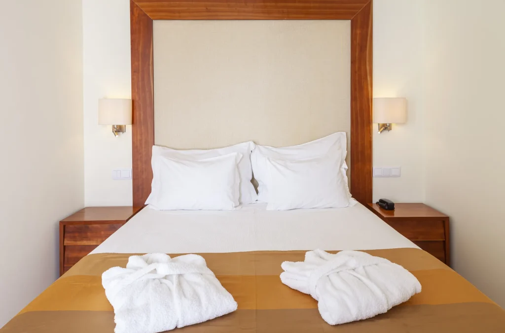Hotel bed with white linens, wooden headboard, two bedside lamps, and neatly folded bathrobes on the duvet.