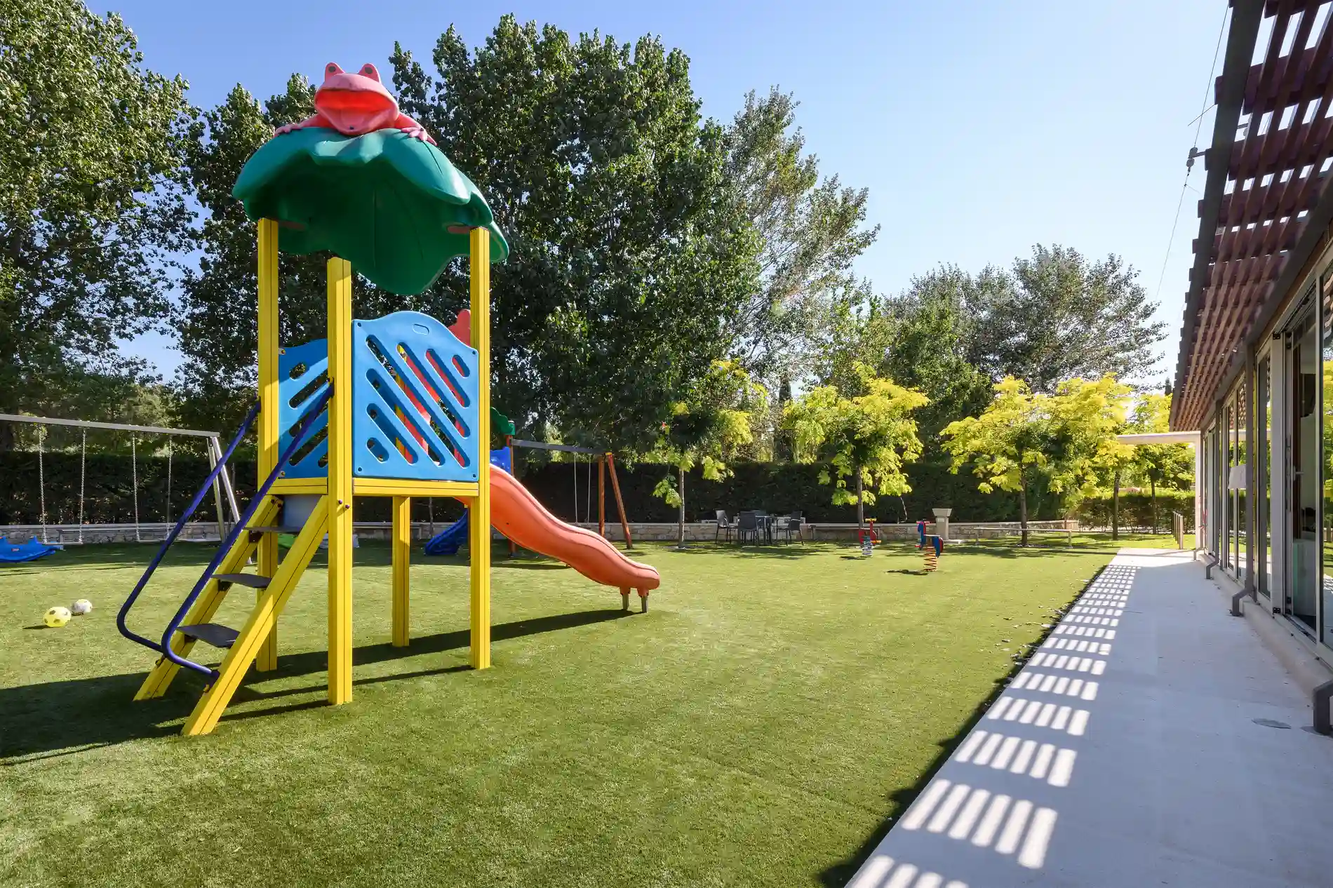 Parque infantil com escorregador e balanço, cercado por árvores, grama verde e espaço aberto sob céu azul claro.