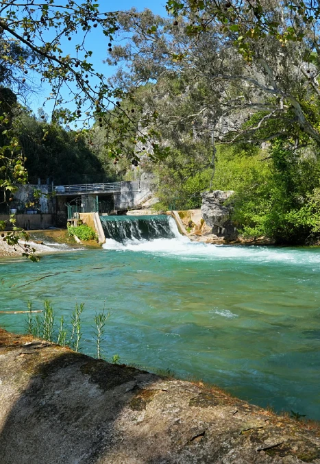 Pequena cachoeira em rio de águas claras, cercada por árvores e vegetação densa sob céu azul.