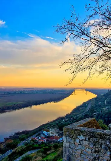 Vista de um rio refletindo o pôr do sol, com árvores e construções em uma colina ao redor.