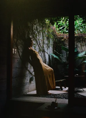 Woman in a sunlit courtyard near a stone wall and greenery, casting shadows
