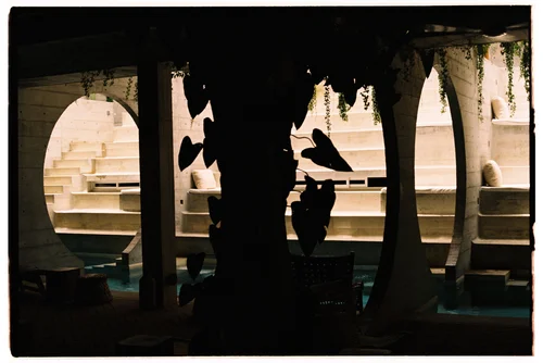 Silhouetted figures in a dim windowed courtyard with arches and benches