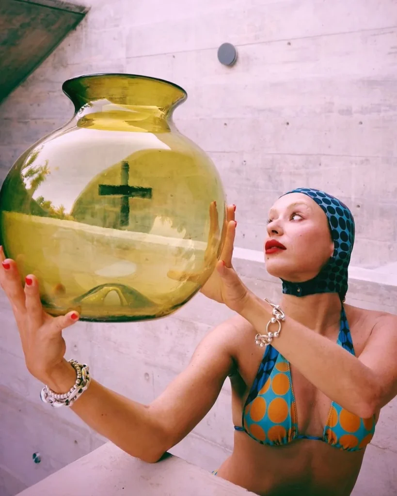 Woman in blue polka-dot bikini holds a glass vase with a green cross inside