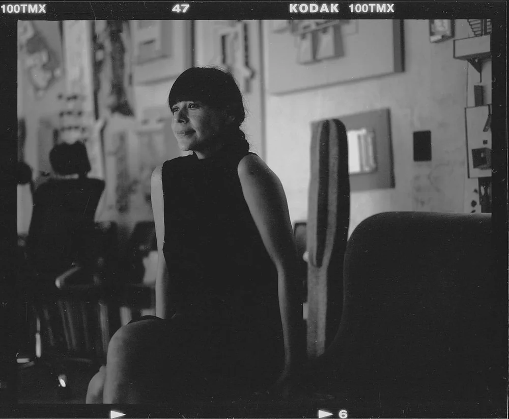 Black-and-white photo of a woman in a black dress sitting indoors.