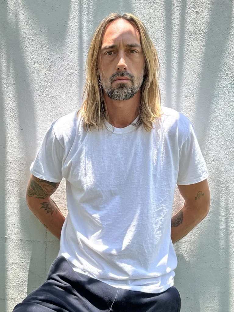 Man with long blond hair and a gray beard wearing a white T-shirt outdoors
