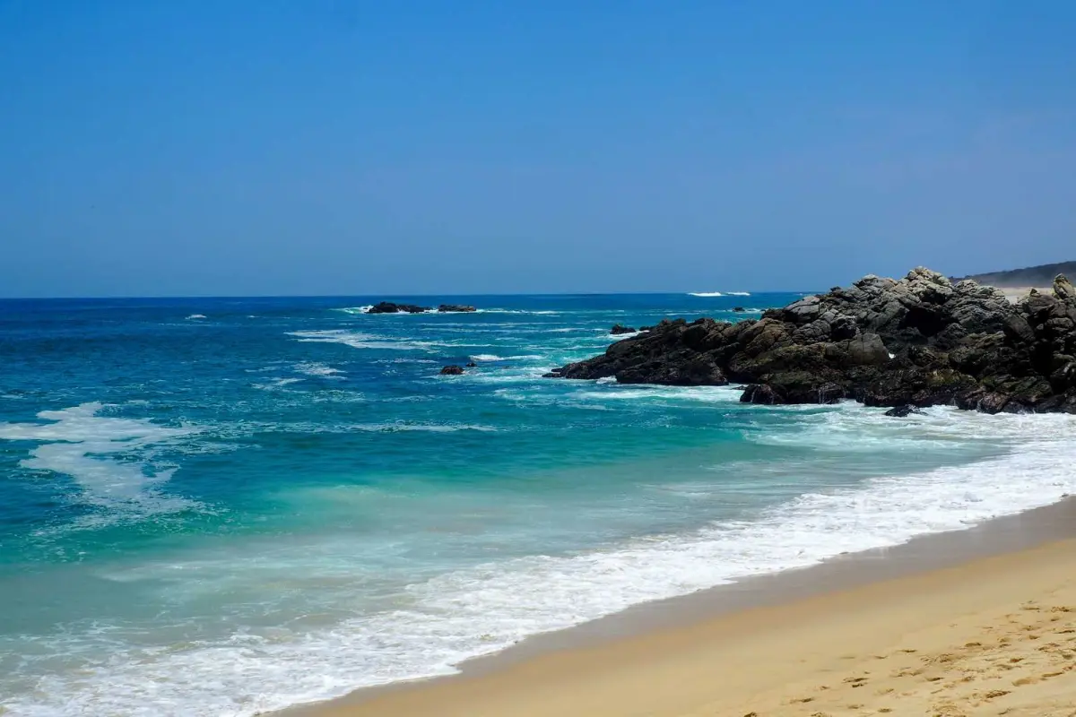 Blue ocean waves and rocky coastline with sandy beach under clear sky