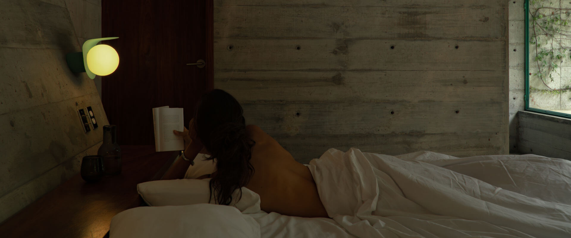 Woman reading a book in bed by a lit wall lamp in a dim room
