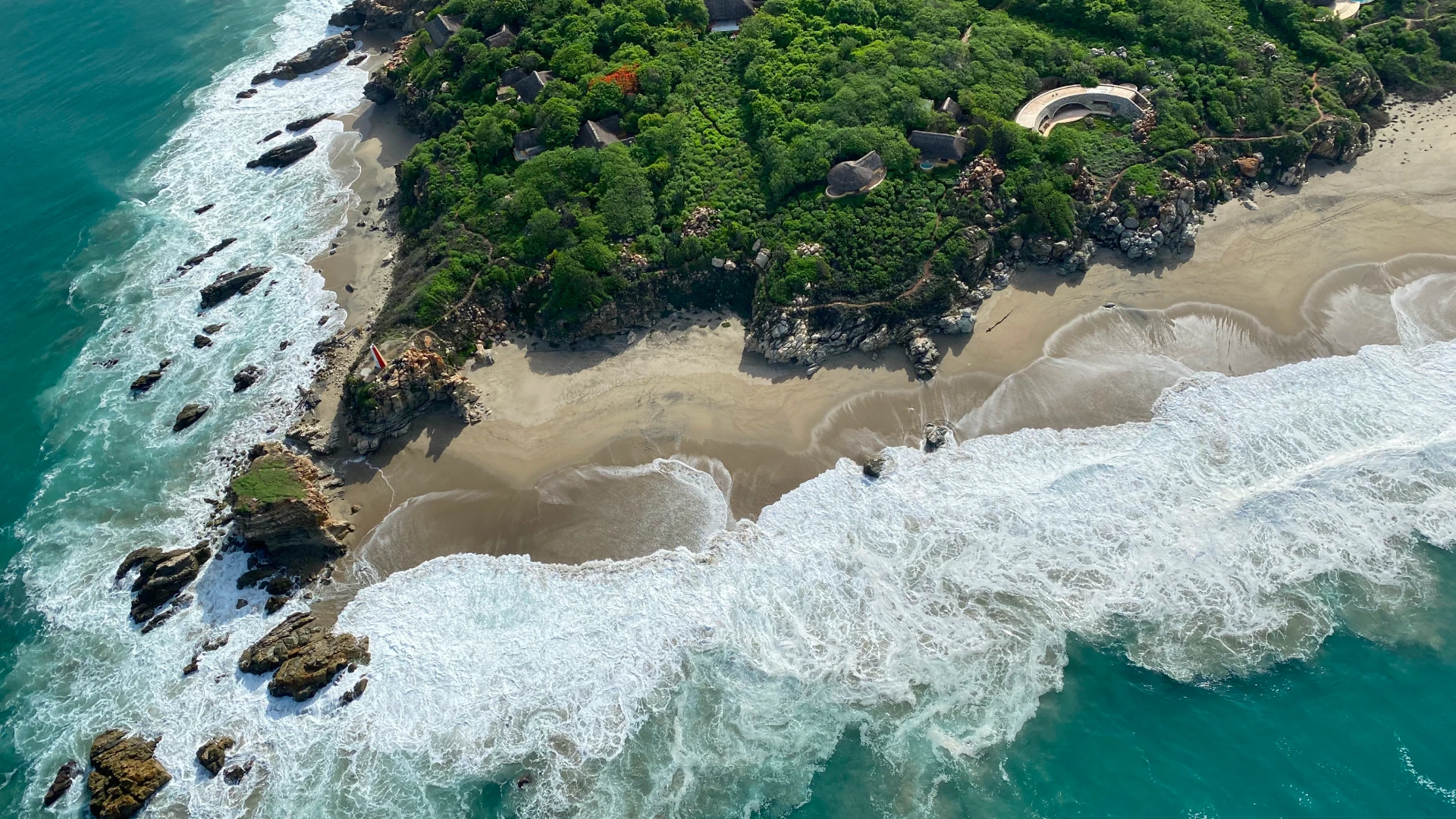 Aerial view of rocky coastline, sandy beach, and waves near green jungle cove