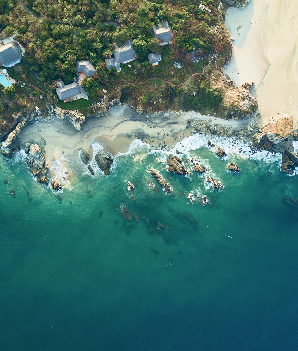 Aerial view of a tropical coastline with houses, waves, and rocky reefs.